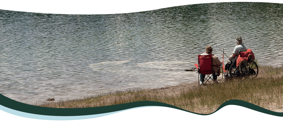 Two people sit and fish on the side of a lake.  One person is in a wheelchair. The other is sitting in a red folding chair. 