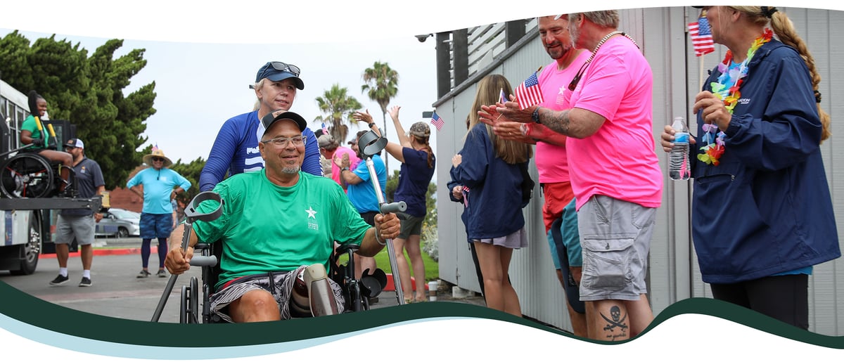 An older man in a green shirt and wheelchair is being pushed down a street by a woman, while people cheer around them.